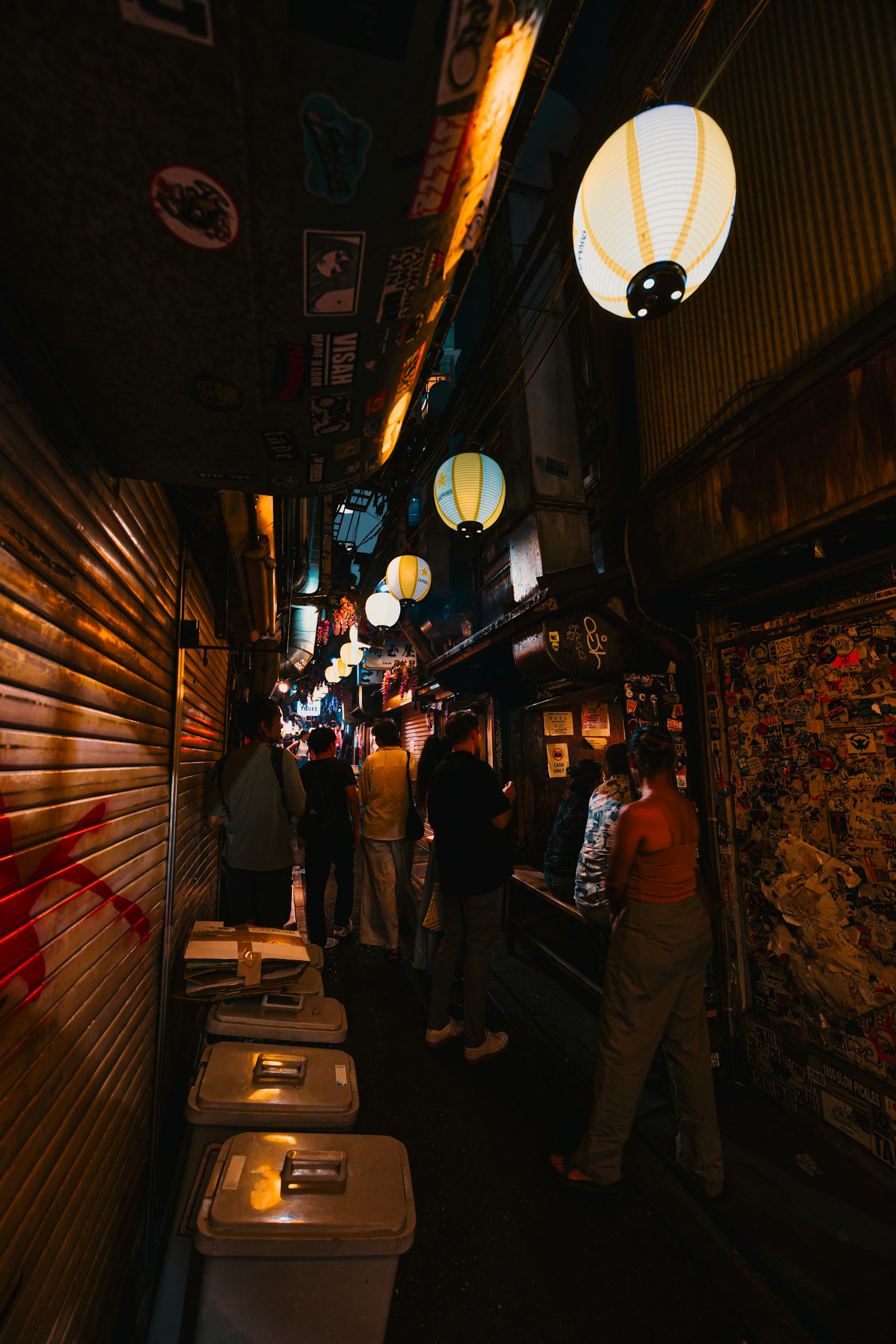 A narrow Japanese alley at night lined with small shops and glowing paper lanterns