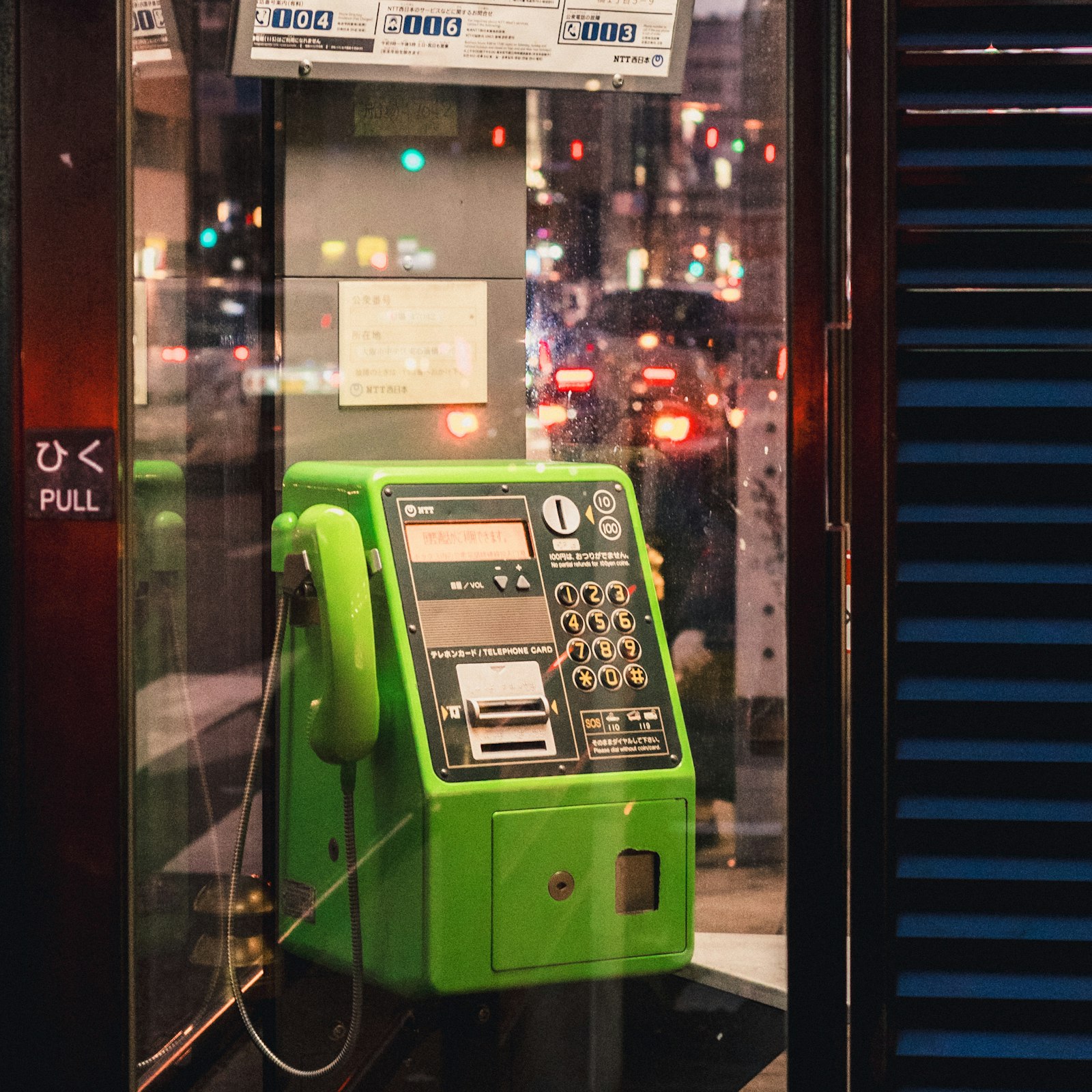 A bright green NTT public payphone in a glass phone booth on a Tokyo street at night